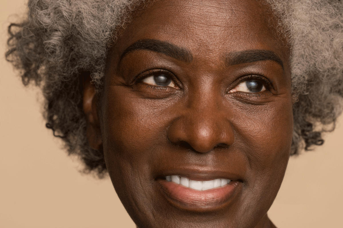 A close up on a black woman's face. She's maybe 50 or 60 years old. Her hair is in a natural style and is grey. She is looking warmly up and to the right,